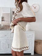 Woman wearing a cream-colored two-piece outfit with brown embroidered floral and wavy patterns, featuring a short-sleeve button-up top and a matching skirt, standing indoors with clear heels and smiling.