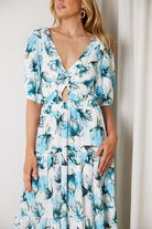 Woman wearing a white and blue floral midi dress with a front cut-out and ruched detailing, featuring elbow-length sleeves and a flowy skirt, standing indoors and smiling.