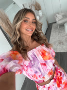 A woman stands indoors in a well-lit, modern room with sheer white curtains and minimalist white cabinetry. She is wearing a colorful off-the-shoulder two-piece outfit consisting of a ruffled crop top and a flowing maxi skirt with a vibrant watercolor floral print in pink, purple, orange, and red hues. Her wavy hair is styled loosely, and she stands gracefully with her hands gently clasped in front, exuding a fresh, summery elegance.