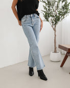 Woman wearing light-wash flared jeans and black heeled ankle boots, paired with a sleeveless black top, standing indoors near a potted plant and a wooden chair.
