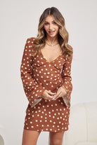 Woman wearing a brown long-sleeve mini dress with white polka dots and flared cuffs, paired with light beige ankle boots, standing in a modern, minimalist room with a white chair, a small shelf, and a textured rug.