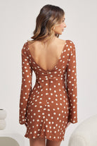 Woman wearing a brown long-sleeve mini dress with white polka dots and flared cuffs, paired with light beige ankle boots, standing in a modern, minimalist room with a white chair, a small shelf, and a textured rug.