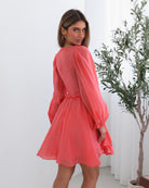 A woman wearing a coral-colored mini dress with a v-neckline, long bishop sleeves, smocked bodice, frills on waist, cut out, and an a-line skirt.