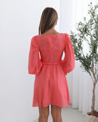 A woman wearing a coral-colored mini dress with a v-neckline, long bishop sleeves, smocked bodice, frills on waist, cut out, and an a-line skirt.