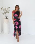 A woman standing in a white room wearing a black floral maxi dress with asymmetric neckline and ruffled skirt.