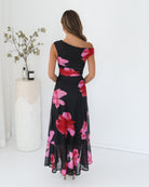 A woman standing in a white room wearing a black floral maxi dress with asymmetric neckline and ruffled skirt.