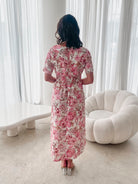 A woman standing in a room with a white chair, wearing a midi dress with a wide v-neckline, short puff sleeves, and an ornamental rose print.