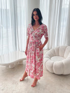 A woman standing in a room with a white chair, wearing a midi dress with a wide v-neckline, short puff sleeves, and an ornamental rose print.