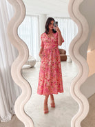 A person sitting on a white sofa wearing a peach blossom midi dress with a wide v-neckline, short puff sleeves, and a flowy a-line skirt.