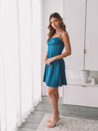A woman standing in a minimalist setting wearing a teal mini dress with slim straps and a sweetheart neckline.