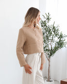 A woman wearing a beige Jacket with long sleeves, round neckline, and center-front buttons. The jacket has a crop length and she is paired with light-colored trousers.