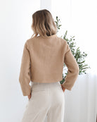 A woman wearing a beige Jacket with long sleeves, round neckline, and center-front buttons. The jacket has a crop length and she is paired with light-colored trousers.