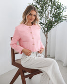 A woman wearing a pink Valdivia Jacket with long sleeves, round neckline, and center-front buttons. The jacket has a crop length and she is paired with light-colored trousers.