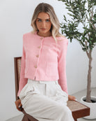 A woman wearing a pink Valdivia Jacket with long sleeves, round neckline, and center-front buttons. The jacket has a crop length and she is paired with light-colored trousers.