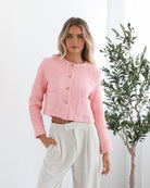 A woman wearing a pink Valdivia Jacket with long sleeves, round neckline, and center-front buttons. The jacket has a crop length and she is paired with light-colored trousers.