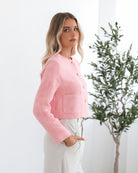 A woman wearing a pink Valdivia Jacket with long sleeves, round neckline, and center-front buttons. The jacket has a crop length and she is paired with light-colored trousers.