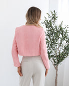 A woman wearing a pink Valdivia Jacket with long sleeves, round neckline, and center-front buttons. The jacket has a crop length and she is paired with light-colored trousers.