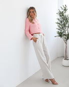 A woman wearing a pink Valdivia Jacket with long sleeves, round neckline, and center-front buttons. The jacket has a crop length and she is paired with light-colored trousers.