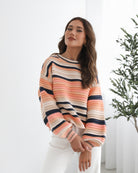 A woman sitting in a chair wearing a striped peach, pink, blue, and black knit top with a round neckline and long sleeves, paired with white pants.