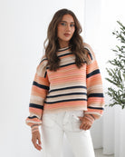 A woman sitting in a chair wearing a striped peach, pink, blue, and black knit top with a round neckline and long sleeves, paired with white pants.