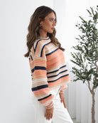 A woman sitting in a chair wearing a striped peach, pink, blue, and black knit top with a round neckline and long sleeves, paired with white pants.