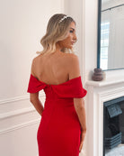 Elegant woman wearing a red off-shoulder gown, perfect for formal events, weddings, evening balls, and special occasions, standing in a sophisticated room with wooden flooring and a fireplace.