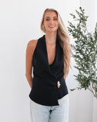 A woman wearing a navy sleeveless top with a plunge neckline and overlap bodice, paired with light jeans. She has blonde hair and is smiling.