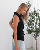 A woman wearing a navy sleeveless top with a plunge neckline and overlap bodice, paired with light jeans. She has blonde hair and is smiling.