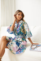 A woman wearing a short, multicolored floral dress with puff sleeves and a belted waist. The dress features a patchwork design with vibrant flower patterns in shades of pink, blue, yellow, and green. She is standing indoors in front of a white couch, looking to the side with a gentle smile.