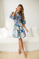 A woman wearing a short, multicolored floral dress with puff sleeves and a belted waist. The dress features a patchwork design with vibrant flower patterns in shades of pink, blue, yellow, and green. She is standing indoors in front of a white couch, looking to the side with a gentle smile.