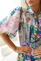 A woman wearing a short, multicolored floral dress with puff sleeves and a belted waist. The dress features a patchwork design with vibrant flower patterns in shades of pink, blue, yellow, and green. She is standing indoors in front of a white couch, looking to the side with a gentle smile.