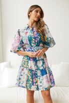 A woman wearing a short, multicolored floral dress with puff sleeves and a belted waist. The dress features a patchwork design with vibrant flower patterns in shades of pink, blue, yellow, and green. She is standing indoors in front of a white couch, looking to the side with a gentle smile.