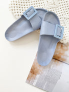 A pair of black summer slides with thick straps, displayed on a flat surface with a knitted texture in the background.