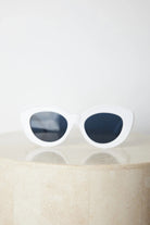 White cat-eye sunglasses with dark lenses displayed on a light beige marble surface against a plain white background.