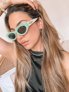 Close-up of a pair of bright green cat-eye sunglasses with dark lenses, placed on a smooth beige surface with a plain white background.