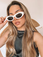 Close-up of a pair of white oval-shaped sunglasses with dark lenses, resting on a smooth beige surface against a plain white background.