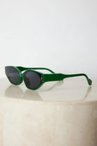 Green geometric-shaped sunglasses with dark lenses resting on a light marble surface against a plain white background.