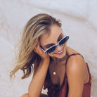 Woman wearing narrow oval-shaped sunglasses with a bold black and white animal print frame, looking over her shoulder with a confident expression. She has wavy blonde hair and is dressed in a rust-colored spaghetti strap top, sitting against a soft beachy backdrop with sand, a light blue door, and a stone wall.