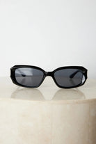 Black rectangular sunglasses with a glossy frame and dark tinted lenses, displayed on a light beige curved surface against a plain white background.