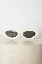 Close-up of a pair of white oval-shaped sunglasses with dark lenses, resting on a smooth beige surface against a plain white background.