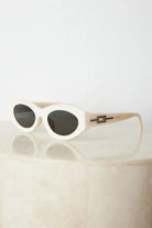 Close-up of a pair of white oval-shaped sunglasses with dark lenses, resting on a smooth beige surface against a plain white background.