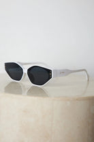 White geometric-shaped sunglasses with dark lenses resting on a light marble surface against a plain white background.