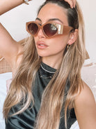 Brown cat-eye sunglasses with thick, glossy frames and dark brown tinted lenses, featuring gold accents on the corners, displayed on a light beige surface against a plain white background.